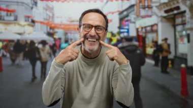 Açık şehir ortamında gülümseyen ve yanaklarını gösteren olgun bir adam. Etrafı bulanık insanlar ve renkli sokak dekorasyonlarıyla çevrili..
