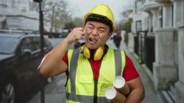 Güvenlik yeleği ve miğferi olan Asyalı genç adam komik bir şekilde şehir sokaklarında hareket ediyor, elinde neşeli bir ifadeyle planlar tutuyor..
