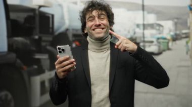Şehir caddesinde gülümseyen genç adam telefonuna işaret ederken, şık giyinmiş, açık havada, canlı bir şehir ortamında, kendine güven ve cazibe yayıyor..