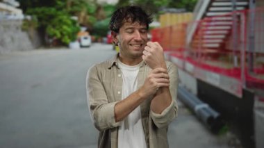 Açık şehir ortamında bileğini tutarken gülümseyen İspanyol bir adam günlük kıyafetlerle acı ve yaralanma ifadesini gösteriyor..