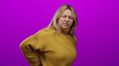 Sarışın kadın, parlak pembe bir duvarın önünde rahat sarı bir kazak giyerek sırt ağrısı çekiyor. Rahatsızlığını dile getiriyor ve izole bir ortamda sağlık ve yaşam tarzı temalarını yakalıyor..