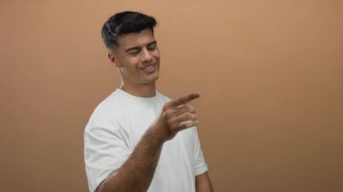 Genç adam rahat bir şekilde gülümsüyor. Yumuşak kahverengi bir arka plana karşı. Çekici ve arkadaş canlısı bir görünüm sergiliyor..