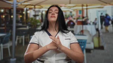 Uzun siyah saçlı genç bir kadın açık hava restoranının terasında oturuyor, elleriyle işaret ediyor, beyaz bir üniforma giyiyor, sıradan bir etkileşime giriyor..