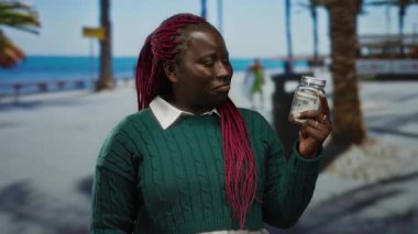 Deniz kenarındaki gezinti güvertesinde elinde dolar banknotuyla cam kavanoz tutan, yeşil kazak giyen, güneşli sahil arka planına karşı duran, kırmızı örgülü ve açık havada okyanusa bakan bir kadın..