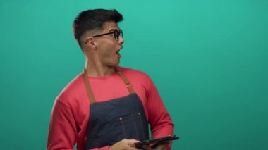 Gözlüklü ve önlüklü genç adam yeşil arka planda tablet tutuyor, düşünceli ve nişanlı görünüyor..