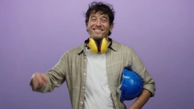 Ses geçirmez kulaklıklar takan İspanyol bir adam ve elinde mor bir arka plana karşı mavi bir baret var. Stüdyo ortamında inşaat ve güvenlik unsurları sergiliyor..