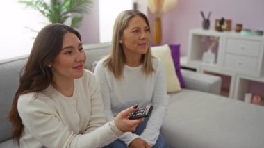 Anne ve kızı sıcak bir oturma odasında uzaktan kumandayla birlikte gülüyorlar, sıcak bir aile anını vurguluyorlar evde rahat eden iki kadınla.
