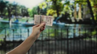Açık hava parkında Suudi riyal banknotları tutan adam arka planda bulanık doğa ve eğlence sahnesini tasvir ederek para ve konumu vurguluyor..