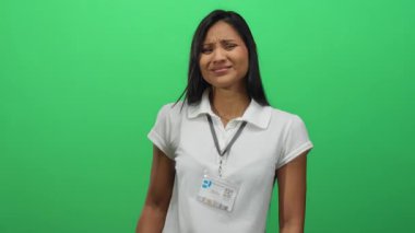 Beyaz tişörtlü, yeşil arka planda el kol hareketi yapan, dışlanmış ve esrarengiz bir duruş sergileyen Latin kökenli genç bir kadın..