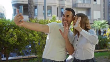 Kadın ve erkek gülümsüyor ve canlı bir parkta selfie çekiyor. Bir şehir ortamında, sevgi, birliktelik ve teknoloji paylaşımı yapıyor..