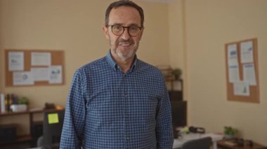 Ofis ortamında gülümseyen, gözlüklü ve rahat gömlekli, profesyonel bir iş yerinde arka planda evraklarla duran olgun İspanyol bir adam..