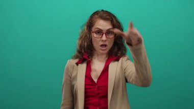 Bir kadın, yeşil duvarın önünde el pençe divan duran, bej ceketli, kırmızı gömlekli, neşeli tavırlı, ezik bir jest yapıyor.