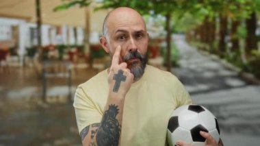 Şehrin bir caddesinde elinde futbol topu olan kel bir adam elinde dövmeler ve göz teması kuruyor..