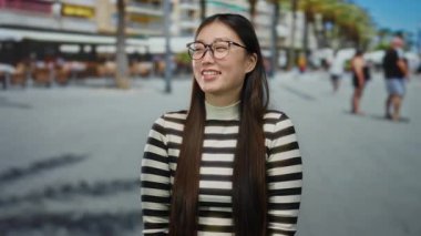 Şehir sokaklarında arka planda gülümseyen bir kadın çizgili gömlek giyen, parlak bir ifadeyle, uzun saç gözlüklü ve neşeli tavırlı bulanık yayalar.