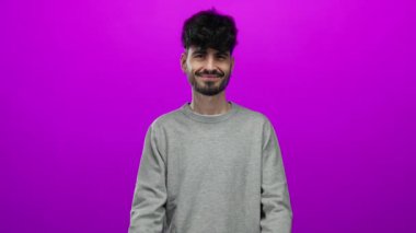 Genç İspanyol bir adam gülümsüyor ve elleri ile işaret ediyor. İzole edilmiş, pembe bir arka planı var. Gri bir süveter giyiyor.