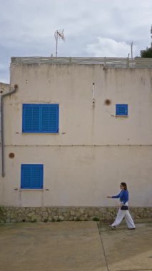 Bulutlu bir günde, geleneksel bir Akdeniz binasının önünden geçerek, eski bir kasabanın Mallorca 'sındaki mavi panjurlarla sahil mimarisini ve yerel kültürü ön plana çıkarıyor..