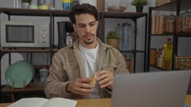 Günlük giysiler içinde genç bir adam, mutfak eşyaları, bir dizüstü bilgisayar ve bir not defteri ile çevrili bir ofiste oturup yemek yiyor, rahat bir çalışma ortamını resmediyor..