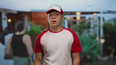 Beyzbol üniformalı genç bir adam parkın dışında, şehir ortamında sıradan ve atletik bir hava sergiliyor..