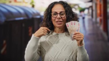 Açık havada dişlerini gösteren, geri dönüşüm kutularının yakınındaki dişlerini gösteren, gözlük ve süveter takan, atık yönetimi ve zenginlik kavramlarını vurgulayan bir kadın..