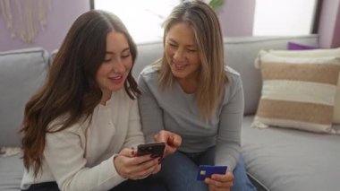 Anne ve kızı rahat bir oturma odasında oturmuş, akıllı telefon kullanırken ve kredi kartı tutarken birlikte kaliteli zaman geçiriyorlar, aile bağlarının ve teknolojinin sıcaklığını gösteriyorlar..