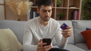 Oturma odasında akıllı telefon ve kredi kartı tutan genç adam, online alışveriş sürecine odaklanmış..
