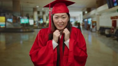 Kırmızı mezuniyet kıyafeti giymiş, havaalanı terminalinde gülümseyen, kadın eğitim ve seyahatlerini ön plana çıkaran, canlı kırmızı elbisesi ve iç mekan ayarlarıyla başarıyı ve yolculuğu vurgulayan bir kadın..