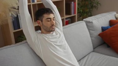 Genç adam rahat bir oturma odasında gözleri kapalı, kolları başının üstünde, kitaplar ve bitkilerle çevrili, huzurlu bir ev ortamı yaratıyor..