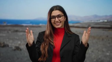 Kırmızı tişörtlü ve siyah ceketli genç İspanyol kadın okyanus manzaralı bir plajda el kol hareketi yaparak kendine güvenen bir hava sergiliyor.