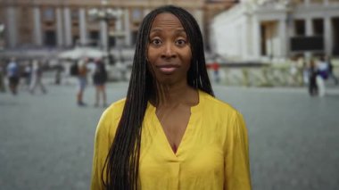 Sarı gömlekli, saçları örgülü bir kadın arkada bulanık yayalar ve tarihi mimariyle çevrili olarak Vatikan meydanında duruyor..