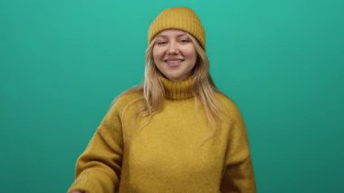 Sarı kazaklı ve şapkalı gülümseyen sarışın kadın yeşil arka planda burnuna dokunuyor..