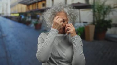 Gri saçlı yaşlı kadın, şehir sokaklarında cep telefonuyla konuşuyor. Endişelerini dile getiriyor ve şehir ortamında modern bağlantıyı ve yaş çeşitliliğini vurguluyor..
