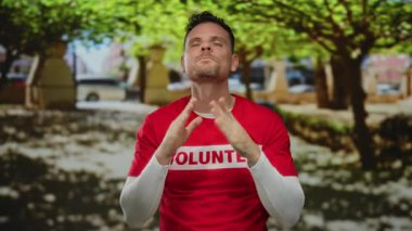 Park ortamında duyguları ifade eden gönüllü tişört giyen, ağaçlar ve güneş ışığıyla çevrili bir adam..