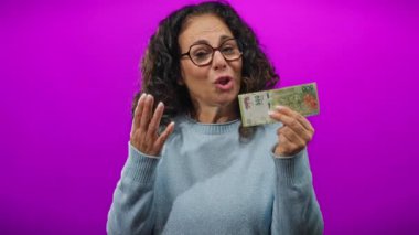 Kafası karışık bir ifadeyle Arjantin pezosu tutan orta yaşlı bir kadın canlı mor bir duvara karşı duruyor ve bu da finans ve kafa karışıklığı temaları öneriyor..