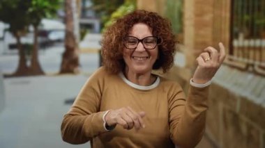 Şehir caddesinde gitar çalan, esmer, orta yaşlı, Latin kökenli, gözlüklü ve kahverengi kazaklı bir kadın, neşeli bir açık hava ifadesi yakalıyor..