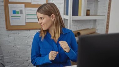 Ofis ortamında gülümseyen ve konuşan bir kadın, duvarında belgeler ve masada bir dizüstü bilgisayarla..