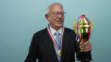 Takım elbiseli kıdemli işadamı elinde bir kupa tutarken mavi bir arkaplana başparmak sallamak zafer ve başarı getirir..