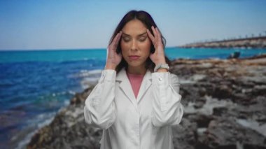 Deniz kenarında gözleri kapalı, elleri şakakların üzerinde beyaz bir ceket giymiş bir kadın okyanus kıyısındaki parlak mavi gökyüzünün altında baş ağrısını yakalıyor..