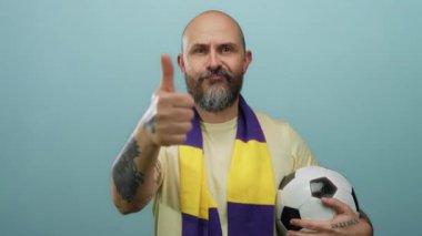 Sakallı, elinde futbol topu olan, mavi arka planda baş parmak gösteren, sarı mor eşarplı, güvenli bir şekilde poz veren beyaz bir adam..