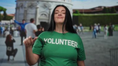 Dışarıdaki kadın kameraya şakacı bir jest yapıyor. Arka planda bir kule var. Gönüllü tişörtü giyiyor..