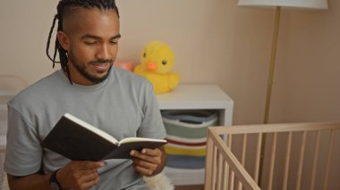 Saçları örgülü genç adam, rahat bir yatak odasında kitap okuyor. Yakınındaki ahşap karyola ve pelüş oyuncakla, sakin ve huzurlu bir ev atmosferi sergiliyor..