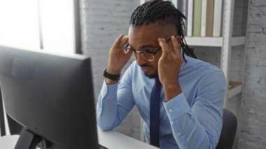 Örgülü genç adam, ofiste mavi bir gömlek giyip, gözlüklerini ayarlarken düşünceli ve profesyonel bir atmosferi yansıtırken endişeli görünüyor..