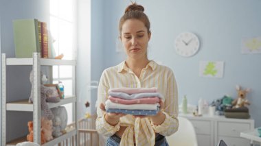 Oyuncak, kitap ve çocuk odası gereksinimleriyle çevrili çamaşırları katlayan kadın, doğal ışık ve huzurlu atmosferle organize ev ortamını yansıtıyor..