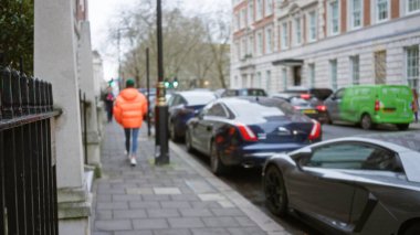 Turuncu ceketli bulanık bir adam kışın Londra caddesinde yürüyor. Etrafı park etmiş arabalar ve tarihi binalarla çevrili. Tipik bir şehir manzarası yakalıyor..