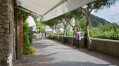 Lüks bir Capri caddesinde, arka planda yemyeşil bir zemin ve taş yapılar arasında yürüyen insanlar, sakin bir ada atmosferini çağrıştırıyor..