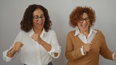 Gözlüklü iki neşeli olgun kadın birlikte gülüyor ve beyaz bir duvar arkasında dans ediyor, canlı bir arkadaşlık ve mutluluk anı yakalıyorlar..