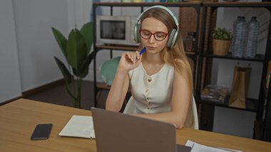 Modern ofiste çalışan bir kadın, dizüstü bilgisayara dikkatlice bakarken kulaklık takıyor. Masasında da raflar ve bitkiler var..