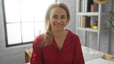 Kapalı bir odada, parlak bir yatak odasında, kırmızı pijamalı, kısa sarı saçlı, kitaplarla çevrili ve rahat ev dekorasyonlu bir evde gülümseyen bir kadın..
