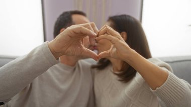 İspanyol çift sıcak bir oturma odasında birlikte oturuyor, elleriyle kalp şekli oluşturuyorlar, evdeki sevgi ve bağlantıyı simgeliyorlar..