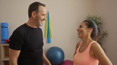 Orta yaşlı İspanyol çift spor salonunda neşeli bir anı paylaşıyor, arka planda egzersiz ekipmanlarıyla aşkı, zindeliği ve arkadaşlığı vurguluyorlar..