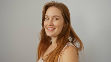 Beyaz bir arka plana karşı uzun kızıl saçlarıyla güvenle gülümseyen bir kadın güzelliğini ve basitliğini gösteriyor..